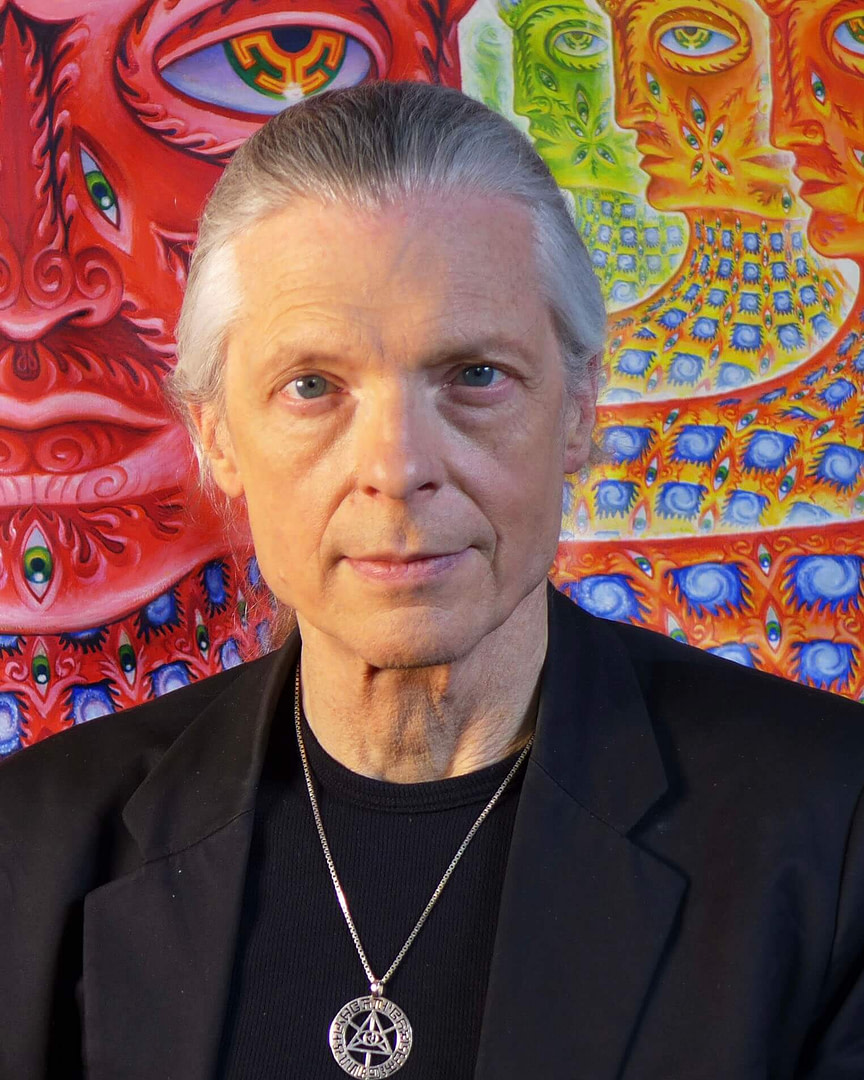 Alex Grey in black standing in front of a colorful painting.