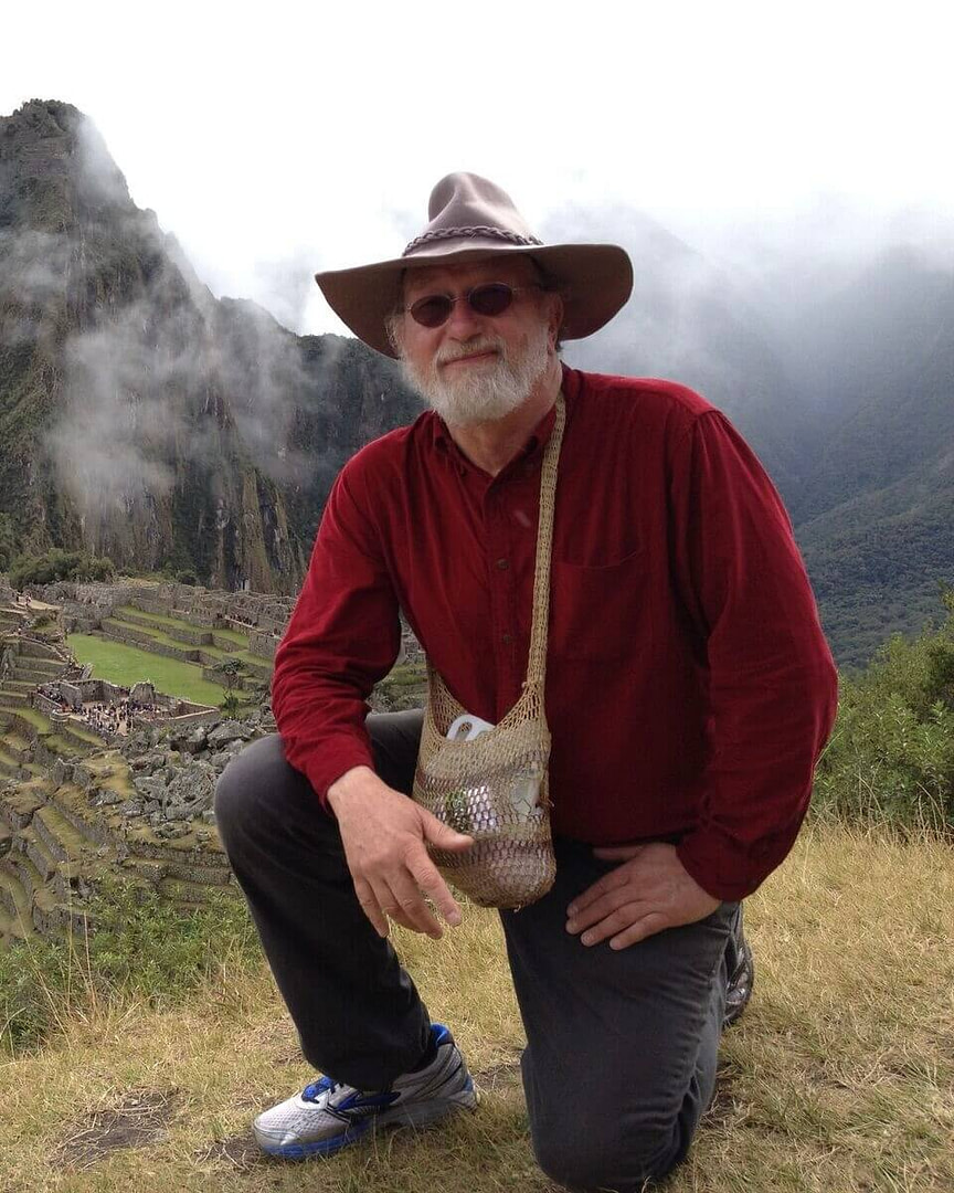 Dr. Dennis McKenna in a hat and a hat squatting on a hill.